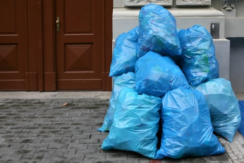 A collection of blue plastic rubbish bags filled with waste materials, stacked and resting on a paved sidewalk area in front of a building with brown wooden doors. The bags are tied at the top and vary slightly in size and shape, with some appearing more bulging and others more tightly packed. The pavement beneath is made of grey bricks, and part of a light-colored wall and a small section of a grey utility box are visible in the background. The image suggests a waste clearance activity, typical of local rubbish removal services, possibly in the Walthamstow area, supporting professional waste management by Office Clearance Walthamstow.