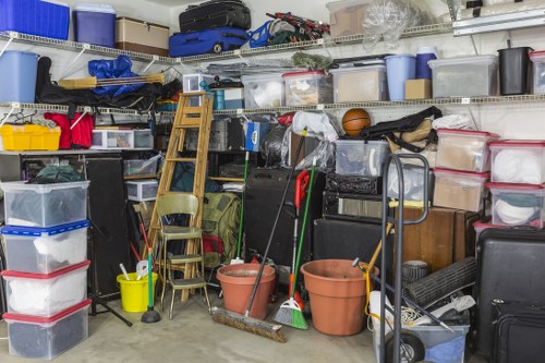 A densely packed indoor storage area or garage filled with a large variety of clutter, including stacked plastic storage bins with red, blue, and transparent lids, some filled with household items, and others closed. Several plastic and cardboard boxes are visible, some placed on metal shelving units extending along the back wall, which also holds suitcases, sports equipment, and miscellaneous containers. In the foreground, there are two large terracotta plant pots, a yellow bucket, and cleaning tools such as brooms and mops leaning against a small wooden chair and a metal trolley. To the left, a step ladder is positioned beside a chair, and additional items like bags and more storage containers are stacked on the floor and on the shelves. The environment appears to be a typical domestic or commercial storage space in Walthamstow, used temporarily for clutter accumulation before rubbish removal or clearance by Office Clearance Walthamstow, set in a well-lit area with visible concrete flooring and a neutral background of shelving and stored goods.