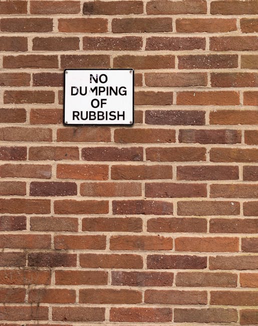 Two large black plastic rubbish bags, filled and tied at the top, are positioned on a pavement next to a curb in an outdoor setting. One bag appears slightly crumpled and leans against a wooden fence
